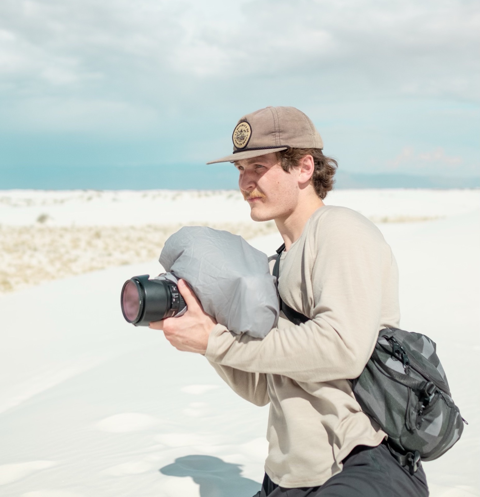 Benjamin holding a camera wrapped in a rain cover on white sand dunes